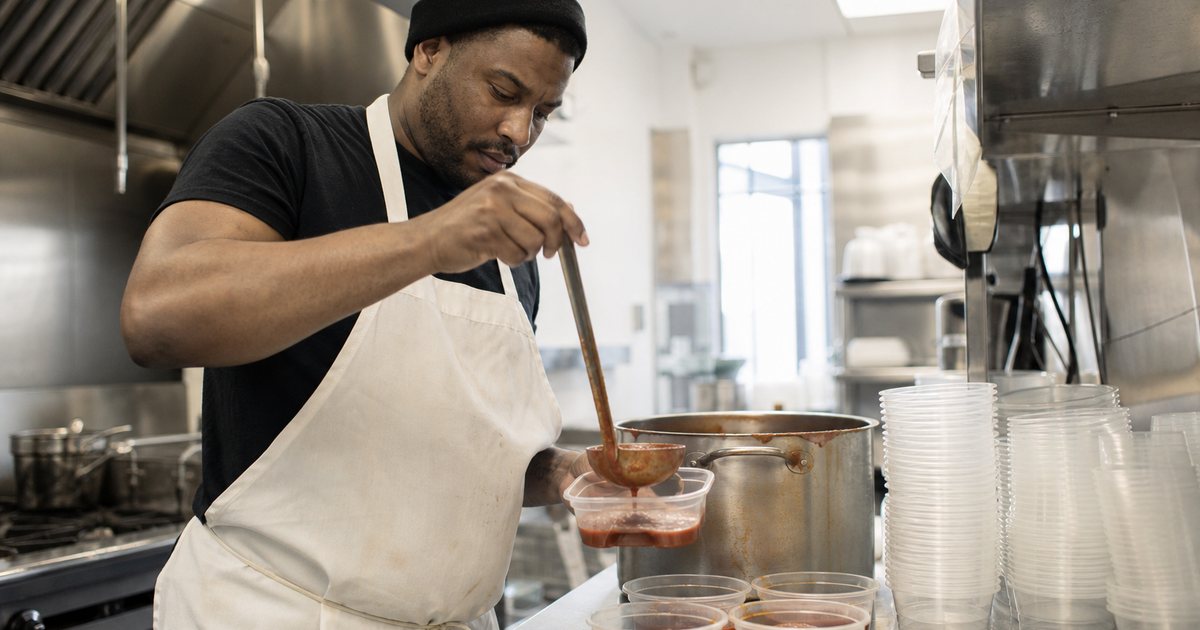 A line cook in a white apron ladling sauce from a large stainless pot into plain clear deli containers on a stainless steel prep table in a bright commercial kitchen, stacks of empty containers beside him, soft morning light from a high window