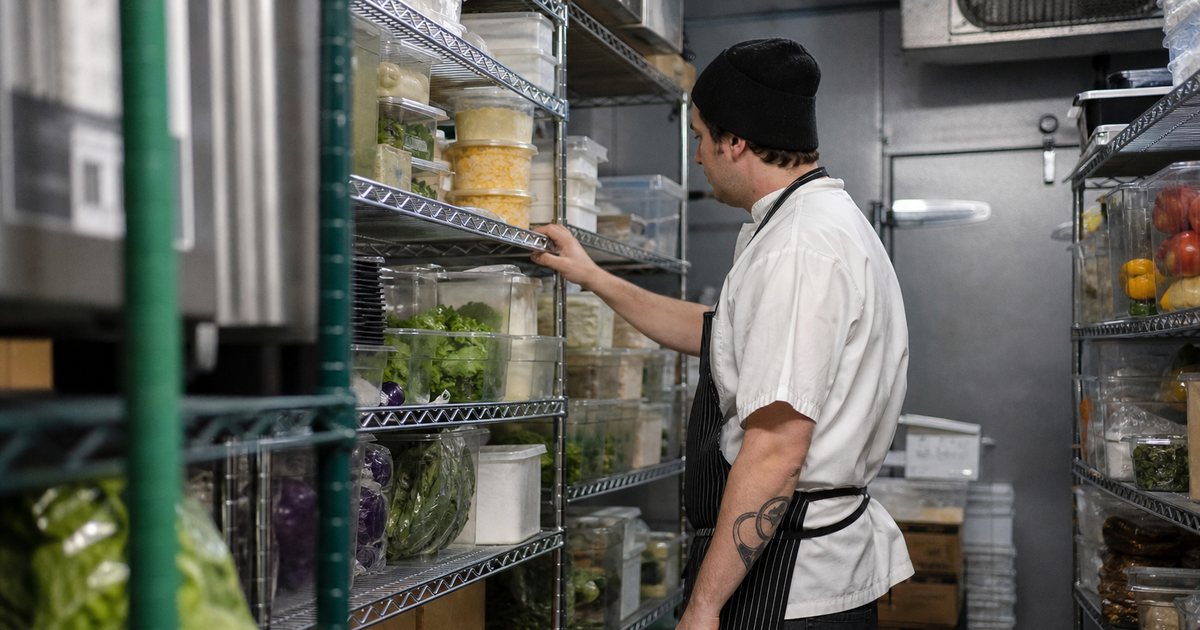 A restaurant walk-in cooler with neatly stacked cases of dairy and produce on metal shelving, one shelf with a few extra cases set aside, a cook in a beanie checking a shelf