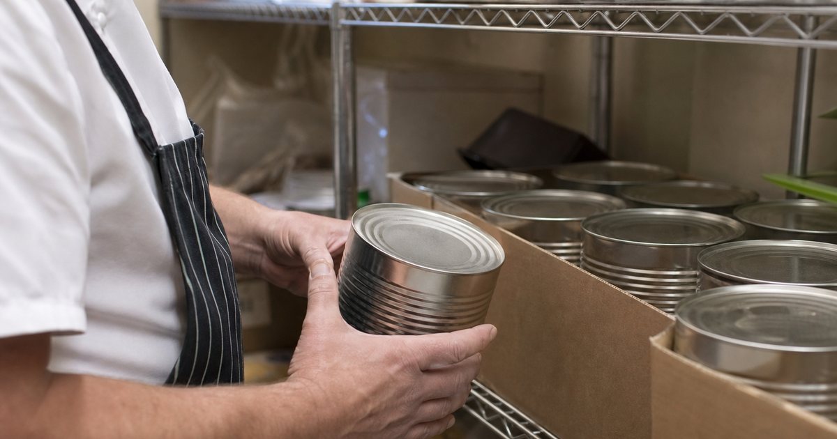 Close-up of a dry storage shelf in a restaurant with a cook holding a large restaurant-size can beside a full cardboard case of the same cans, metal shelving, fluorescent light